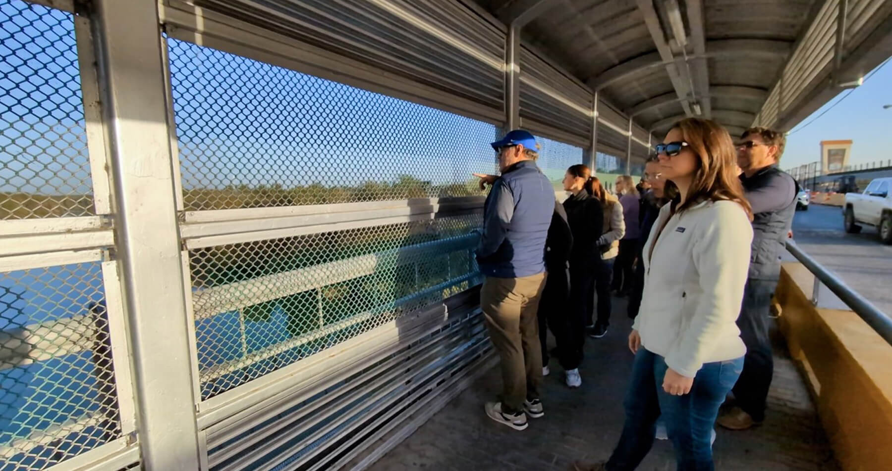 Dr. Venta, wearing sunglasses, stands on an enclosed bridge with others looking through the metal grate at the U.S. border between Texas and Mexico