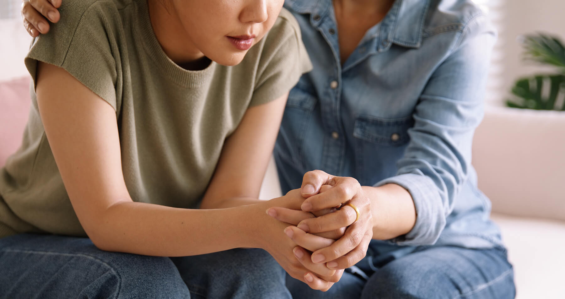 A young woman sits with her arms on her knees, hands clasped. An older woman sits beside her, 1 arm around her shoulder, the other holding her hands