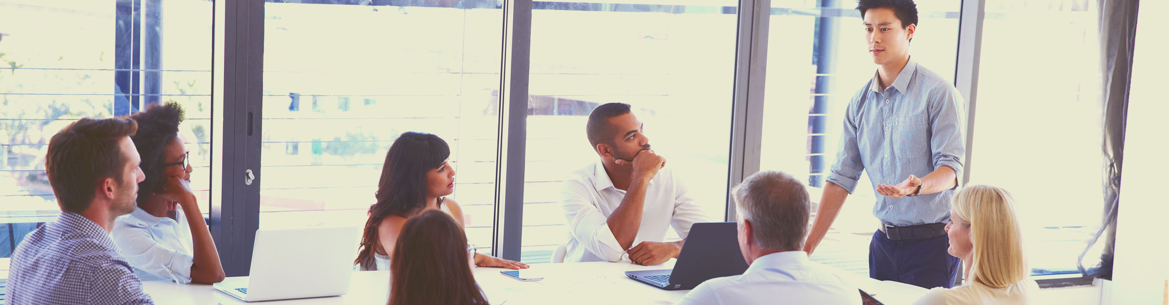 People of different genders, races and ethnicities in a meeting, seated, listening to a man standing at the head of the conference table