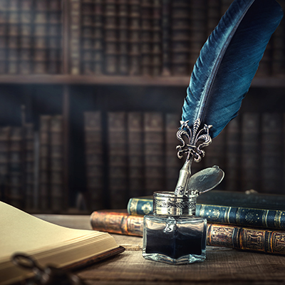 Old quill pen, books and vintage inkwell on wooden desk in the old office against the background of the bookcase and the rays of light.